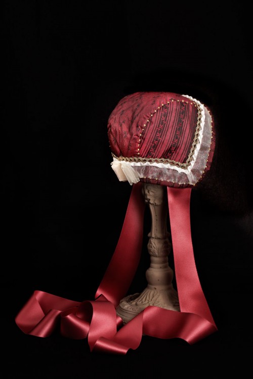 Röd hätta i siden kantad med chiffong och guldspets, dekorerad med chiffongrosett och franska knutar i guldtråd. (Foto Lasse Modin)